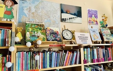 Regał z książkami dla dzieci, nad nim na ścianie kolorowe plakaty, mapa Europy i zegar