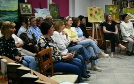 Grupa ludzi siedzi, wok&oacute;ł regały z książkami. 