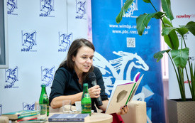 Przy stoliku kobieta z książką i mikrofonem w ręku