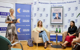 Kobieta wita zebranych gości. Obok przy stoliku prowadząca i gość spotkania