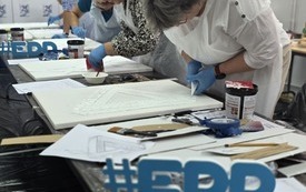 Kobiety podczas nanoszenia gładzi szpachlowej na podobrazie. Na stoliku napisy #EDD oraz materiały wykorzystywane podczas warsztatów.