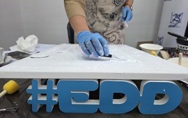 Kobieta nanosi gładź szpachlową na podobrazie. Podobrazie oparte o napis #EDD.