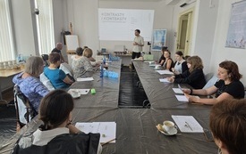 Przy stoliku przykrytym czarną folią siedzi grupa kobiet biorących udział w warsztatach. Obok prowadzący spotkanie wyświetla prezentacje multimedialną na ścianie.