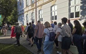Grupa ludzi idących chodnikiem za kobietą w r&oacute;żowej sukience z mikrofonem