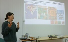 Na ścianie wyświetlana jest prezentacja multimedialna. Obok stoi prowadząca spotkanie kobieta.