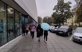 Grupa ludzi z parasolami idzie chodnikiem wzdłuż przeszklonej ściany budynku