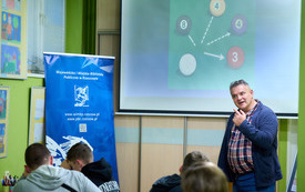 Prowadzący spotkanie mężczyzna mówi do zgromadzonej publiczności. W tle prezentacja multimedialna