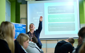 Prowadzący spotkanie mężczyzna z podniesioną do góry ręką mówi do zgromadzonej publiczności. W tle prezentacja multimedialna.
