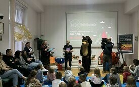 Grupa dzieci z opiekunami. Przed nimi pluszowy miś, dw&oacute;ch mężczyzn, jeden z kamerą i kobieta z mikrofonem