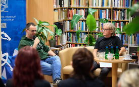 Dwaj mężczyźni we wnętrzu bibliotecznym rozmawiają o poezji. 