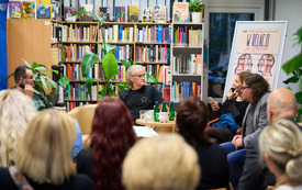 Regały z książkami, kwiaty doniczkowe, uczestnicy panelu poetyckiego. 