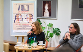 Poetka i poeta uśmiechają się podczas rozmowy. W tle plakat informujący o spotkaniu. 