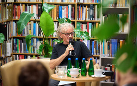 Mężczyzna w dredach wypowiada się do mikrofonu. W tle regały z książkami. 