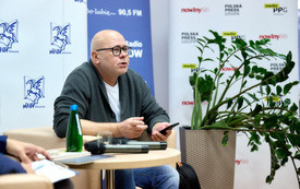 Łysy męzczyzna w okularach z telefonem w ręku opowiada cos publiczności. 