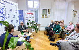 Rozmowa dw&oacute;ch mężczyzn, po prawej zebrana publiczność. 