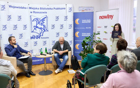 Pisarz, prowadzący spotkanie oraz bibliotekarka są zwr&oacute;ceni twrzą do publiczności. 