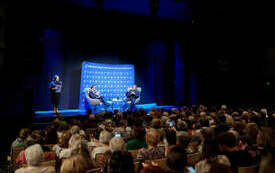W sali teatralnej zebrana widownia. Na scenie pisarz, prowadzący i dyrektorka biblioteki. 