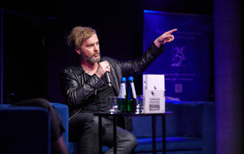 Na fotelu mężczyzna w czarnej sk&oacute;rze i podkoszulku z mikrofonem w ręku.