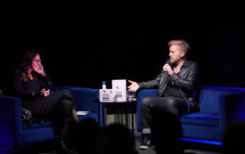 Przy stoliku mężczyzna w czarnej sk&oacute;rze i podkoszulku z mikrofonem w ręku i prowadząca spotkanie kobieta.