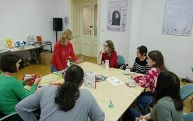 Przy stoliku siedzi grupa os&oacute;b. Obok stolika kobieta w czarownej koszuli.