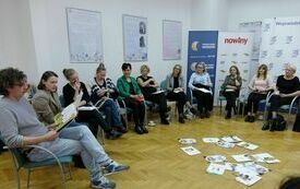 Na krzesłach wok&oacute;ł sali siedzą uczestnicy spotkania. Na podłodze porozrzucane kartki