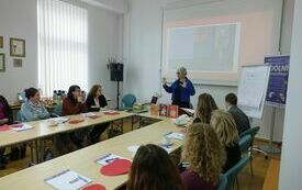 Uczestnicy warsztat&oacute;w siedzą przy stole. Przed nimi prowadząca spotkanie kobieta.