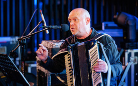 Zdjęcie łysego mężczyzny z akordeonem śpiewającego do mikrofonu. 