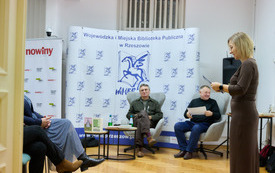 Kobieta stoi i czyta z kartki, w tle za nią siedzą autor i prowadzący spotkanie