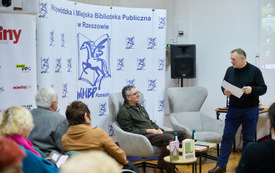 Grupa os&oacute;b na spotkaniu w bibliotece, w tle autor i prowadzący spotkanie