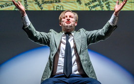 Meżczyzna siedzi na scenie i wznosi ręce ku g&oacute;rze