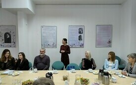 Wśr&oacute;d siedzących os&oacute;b, jedna kobieta przemawia stojąc. 