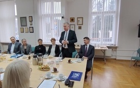 Stojący siwowłosy mężczyzna w garniturze wśr&oacute;d zebranych przy stole konferencyjnym. 