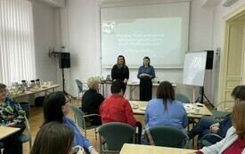 Przy stolikach siedzą uczestnicy spotkania, przed nimi stoją dwie kobiety, w tle prezentacja multimedialna