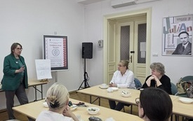 Uczestnicy spotkania słuchają prowadzącej spotkanie kobiety.