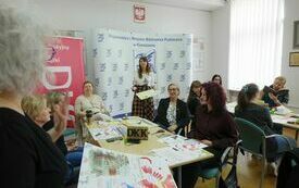 Uczestnicy warsztat&oacute;w siedzą przy stołach, przed nimi prowadząca spotkanie kobieta. 