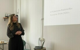 Kobieta w ciemnym ubraniu stoi przy ścianie na kt&oacute;rej wyświetla się prezentacja multimedialna. 