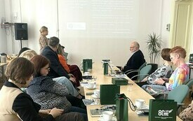 Uczestnicy narady siedzą przy stole, przed nimi kobieta w okularach.