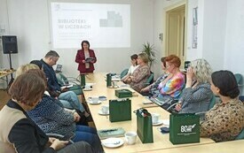 Kobieta w okularach i ciemnych włosach przemawia do zebranych na spotkaniu os&oacute;b.