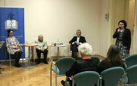 Dwóch mężczyzn i trzy kobiety siedzą na krzesłach, czwarta kobieta stoi i przemawia