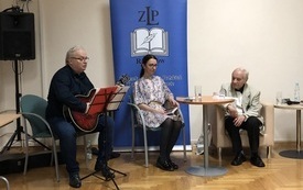 Dwóch mężczyzn siedzi na krzesłach, jeden z gitarą, pomiędzy nimi kobieta