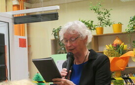 Kobieta w siwych włosach, niebieskiej sukience i okularach, czyta do mikrofonu.