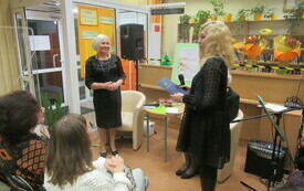 Dwie kobiety na pierwszym planie stoją. W lewym dolnym roku dwie kobiety siedzą na widowni.