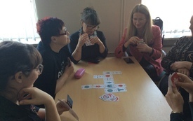 Sześć kobiet siedzi przy stoliku na środku, kt&oacute;rego znajdują się karty do gry. Kobiety trzymają w ręku karty. 
