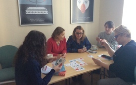 Pięć kobiet siedzi przy stoliku trzymając w ręku karty do gry. Na stoliku rozłożone karty gry.