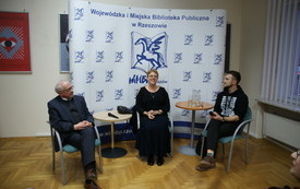 Trzy siedzące osoby na tle baneru Biblioteki. Kobieta siedząca pomiędzy dwoma mężczyznami uśmiecha się. 