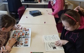 Dwie dziewczynki przeglądają książki z obrazkami, siedząc przy stole. Trzecia dziewczynka stoi przy stole. W tle dwa monitory komputerów.