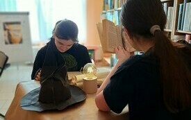 Dwie młode osoby siedzące przy stoliku czytające książki. Na stoliku lampka, zwoje papieru, czapka czarnoksiężnika.