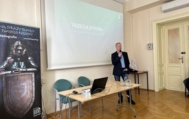 Mężczyzna w marynarce z mikrofonem w ręku stojący przed ekranem projekcyjnym. Obok stolik z laptopem i wodą. 