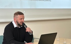 Mężczyzna z brodą trzymający w ręku mikrofon. Przed min ekran laptopa.