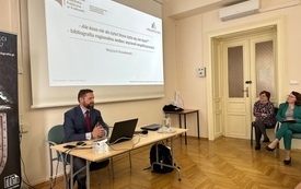 Mężczyzna w średnim wieku z brodą i zarostem siedzący z mikrofonem w ręku przed ekranem laptopa. W tle prezentacja z na ekranie projekcyjnym.